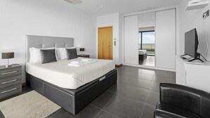 A modern bedroom with a large bed, a bedside table, a TV, and a sliding door leading to a balcony with a pool view. - A&M House - Luxury Holidays Home (Arco da Calheta)
