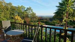 A balcony with a table and chairs overlooking a swimming pool, surrounded by lush greenery and trees.
