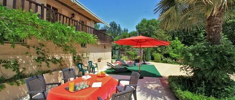 Un patio al aire libre con una mesa preparada para dos personas, rodeado de sillas, un paraguas rojo y abundante vegetación.