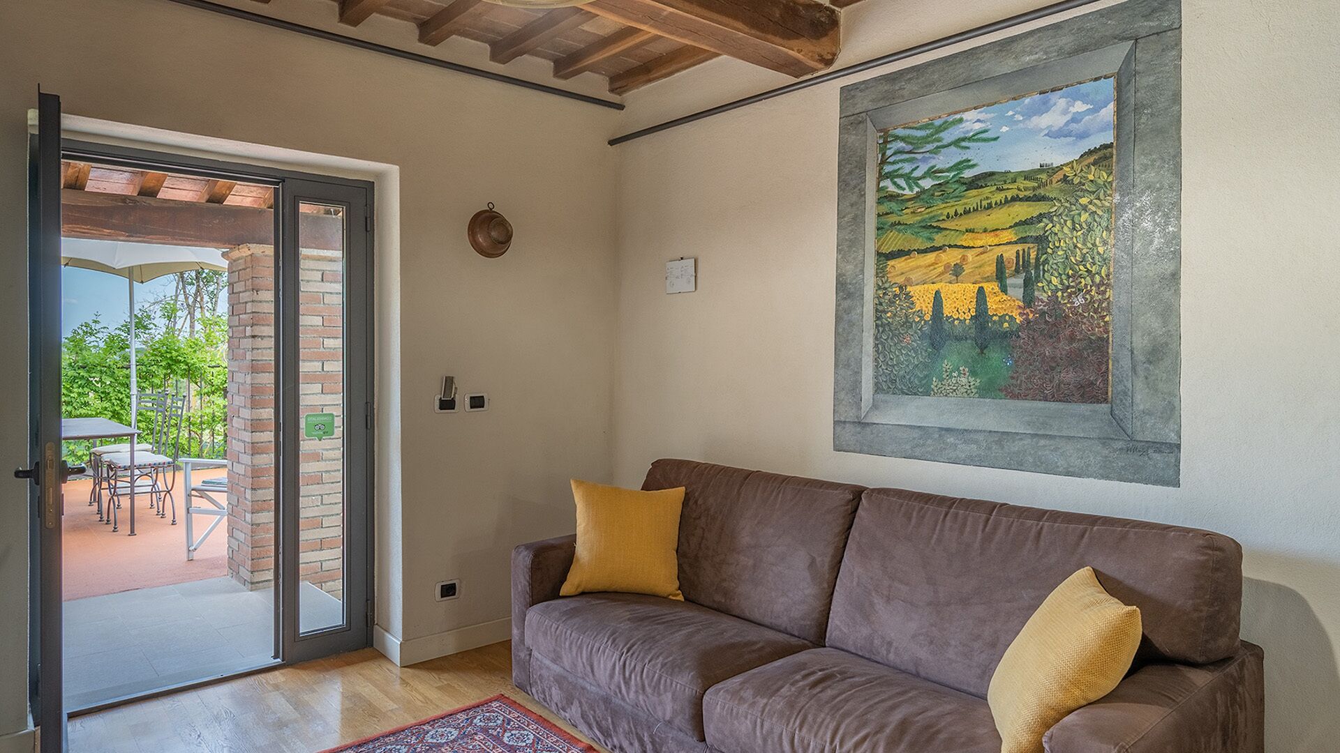 Una habitación con un sofá marrón, un cuadro de un paisaje y una puerta que da a un patio exterior con una mesa y sillas.