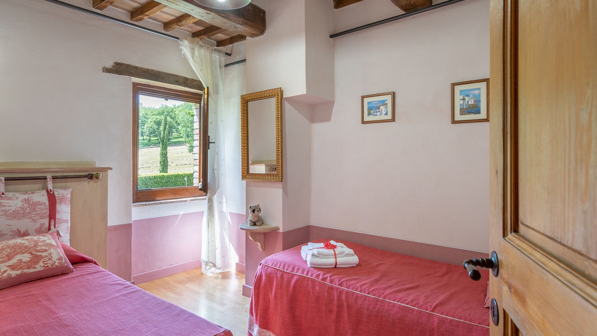 Un dormitorio con dos camas, una ventana con vistas a zonas verdes, un espejo y un estante pequeño con un oso de peluche.