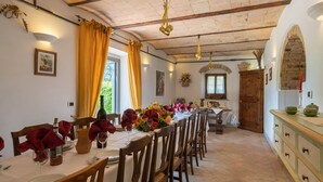 A dining area with a long table set for a meal, wooden chairs, and a kitchen in the background. - La Colonica 18, Emma Villas (Citerna)