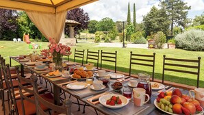 An outdoor dining area with a table set for breakfast, surrounded by chairs, and overlooking a garden with trees and a gazebo. - La Colonica 18, Emma Villas (Citerna)