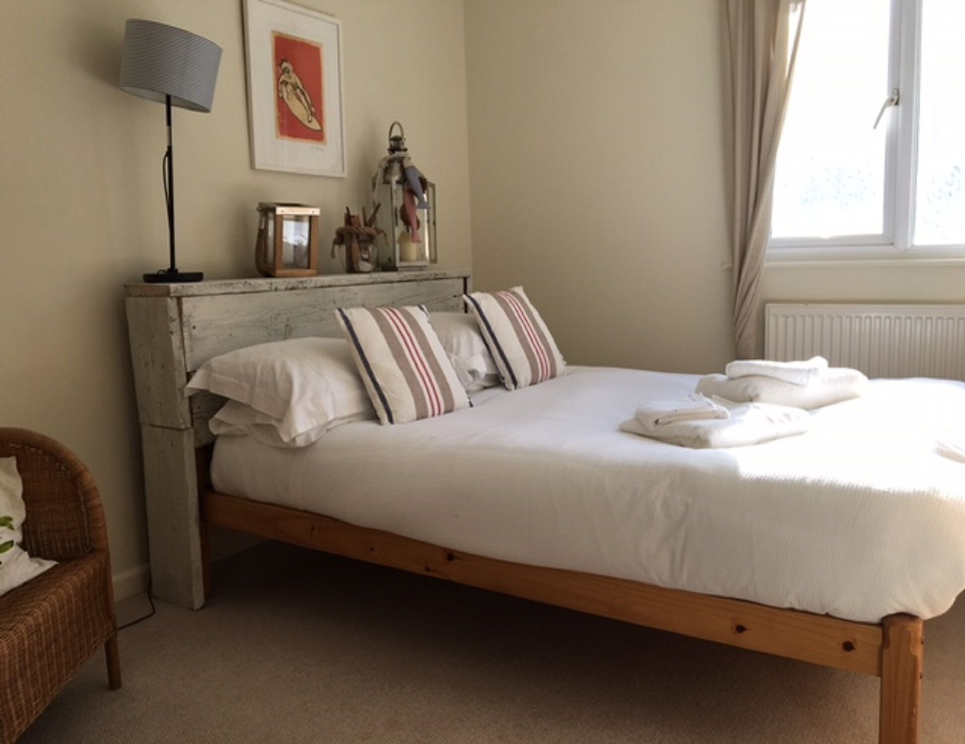 A bed with white bedding, striped pillows, and a wooden frame. There is a lamp, a framed picture, and a window with curtains.