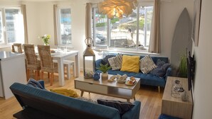 A modern living room with a blue sofa, dining table, and chairs, a coffee table with decorative items, and large windows with a view of the street.