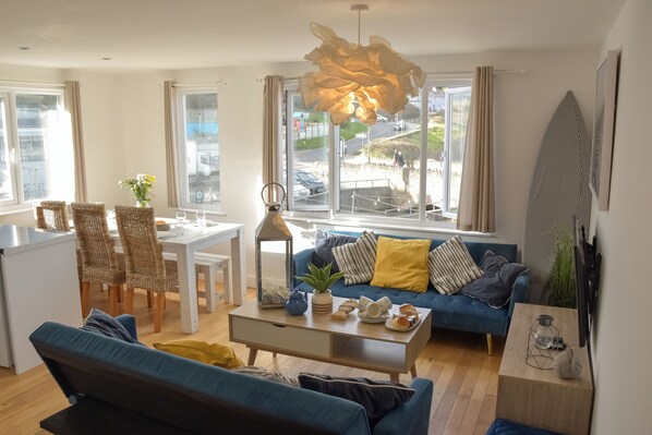 A modern living room with a blue sofa, dining table, and chairs, a coffee table with decorative items, and large windows with a view of the street.