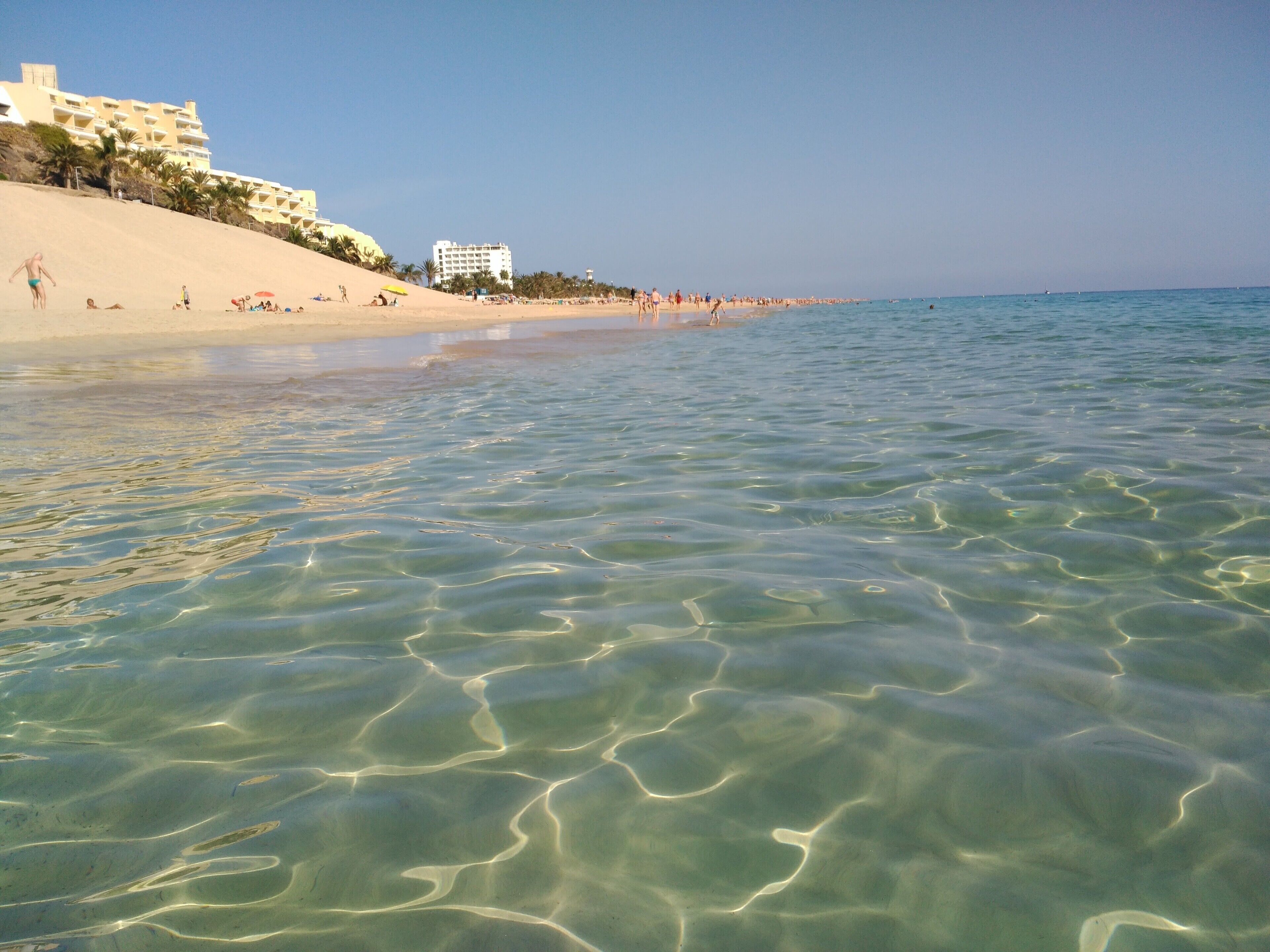 A beach with clear shallow waters, a sandy shore, and buildings in the background.