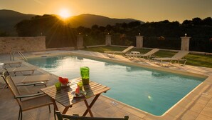 Zona de piscina con sillones de descanso, una mesa con sandía y bebidas, y vista al atardecer sobre las colinas.