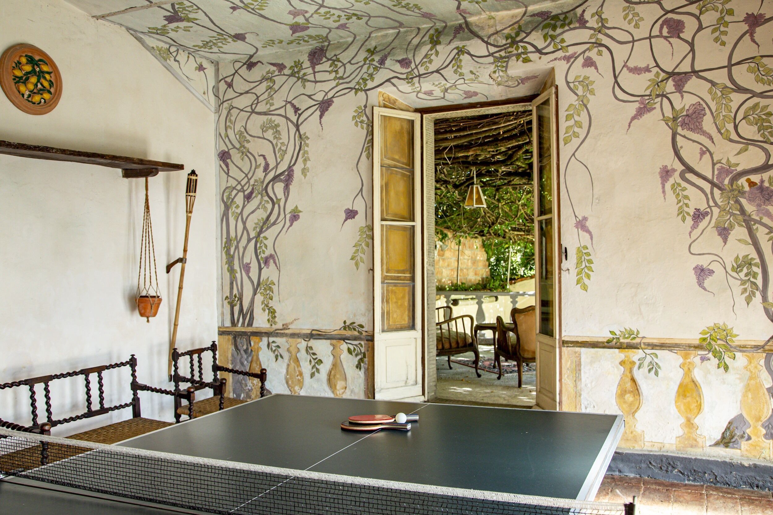 Une table de ping-pong dans une pièce dont un mur est décoré d’une fresque florale, avec un banc en bois et une lampe suspendue.
