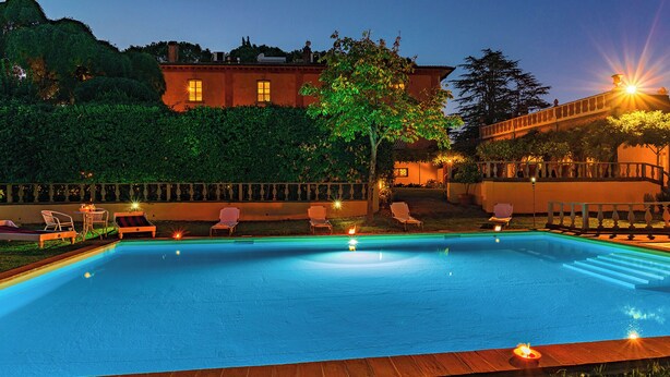 Une piscine rectangulaire bordée d’une terrasse en bois, entourée de chaises longues et d’un espace extérieur bien éclairé, avec un bâtiment visible à l’arrière-plan.