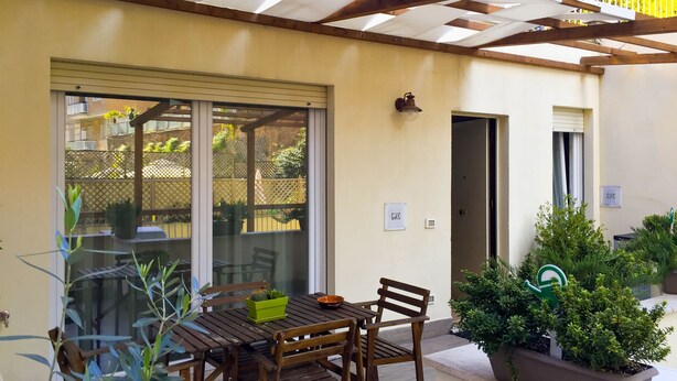 A covered patio with a wooden table and chairs, potted plants, and a sliding glass door leading to a garden. - Elegant house with veranda - in the center of Rome, a stone's throw from S. Giovanni  (Roma)