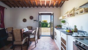 A kitchen with a dining area, wooden ceiling, open door to a garden, and a sink with fruit.