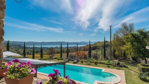 Piscina con vistas a un lago y montañas, rodeada de mobiliario de jardín y plantas en macetas.