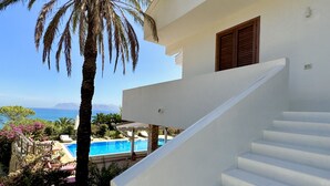 Una scala bianca che conduce a una zona piscina, con una palma e vista sul mare.