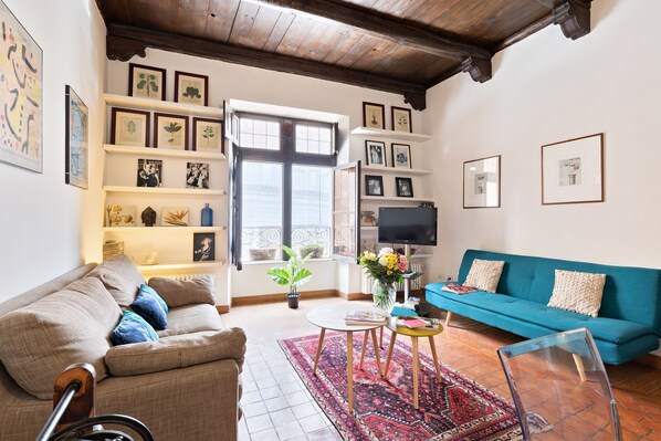 A living room with a sofa, a coffee table, and a TV. There are shelves with framed pictures and a potted plant.