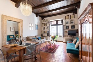 A cozy living room with a wooden dining table, a sofa, and a TV. There are framed pictures on the walls and a chandelier hanging from the ceiling.