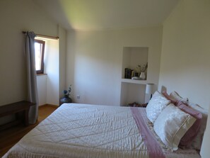A bedroom with a large bed, a bedside table, a nightstand, a window with curtains, a shelf with books and a vase, and a wall-mounted lamp.