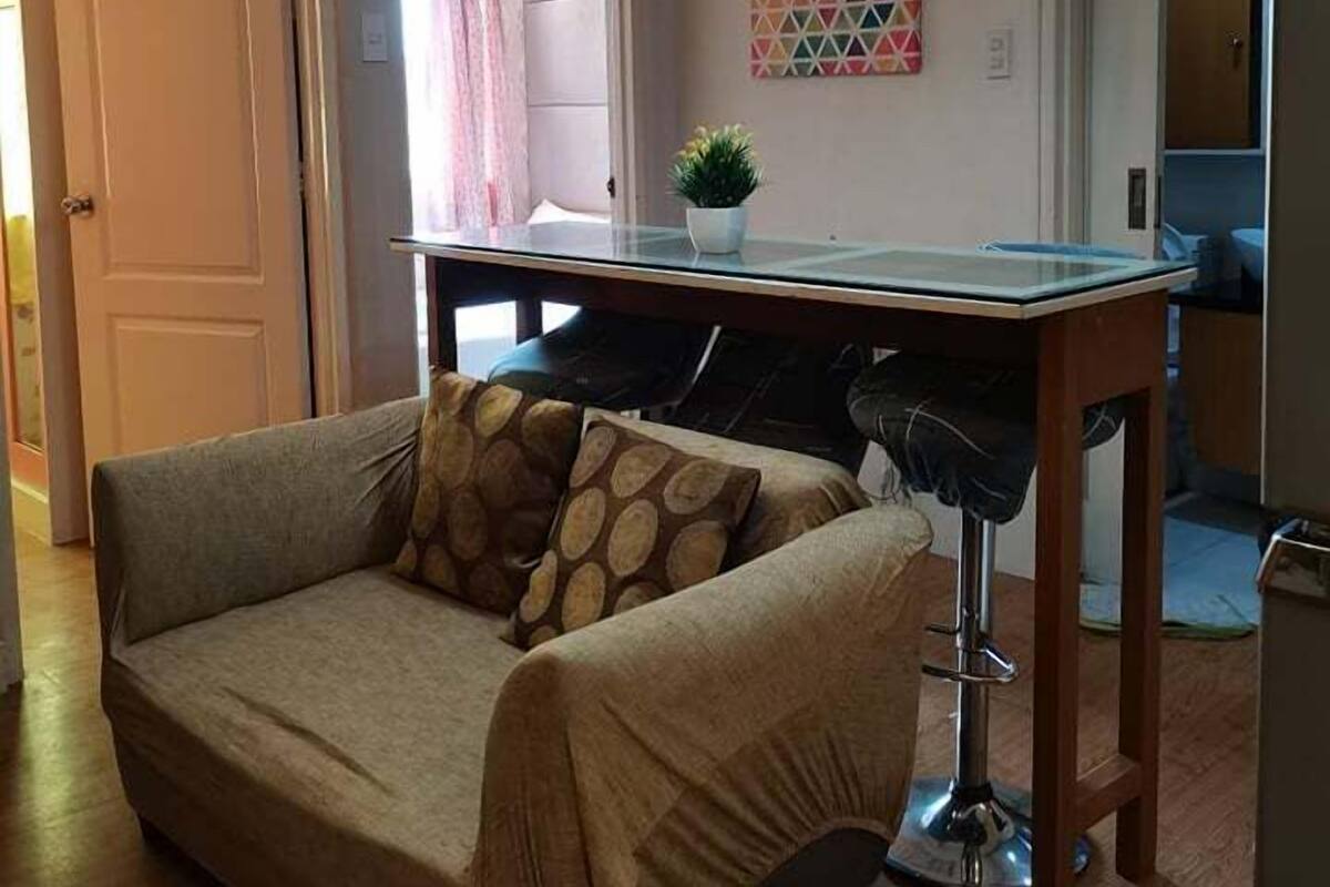 A living room with a sofa, a small table with a plant, and a kitchen area in the background.