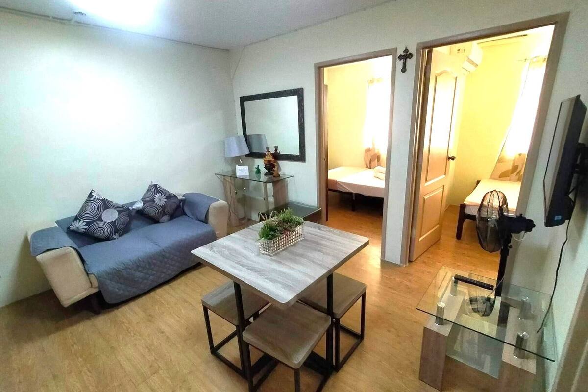 A modern living room with a sofa, a coffee table, and a TV. There is a glass table with a lamp and a small plant. The room has wooden flooring and a door leading to another room.