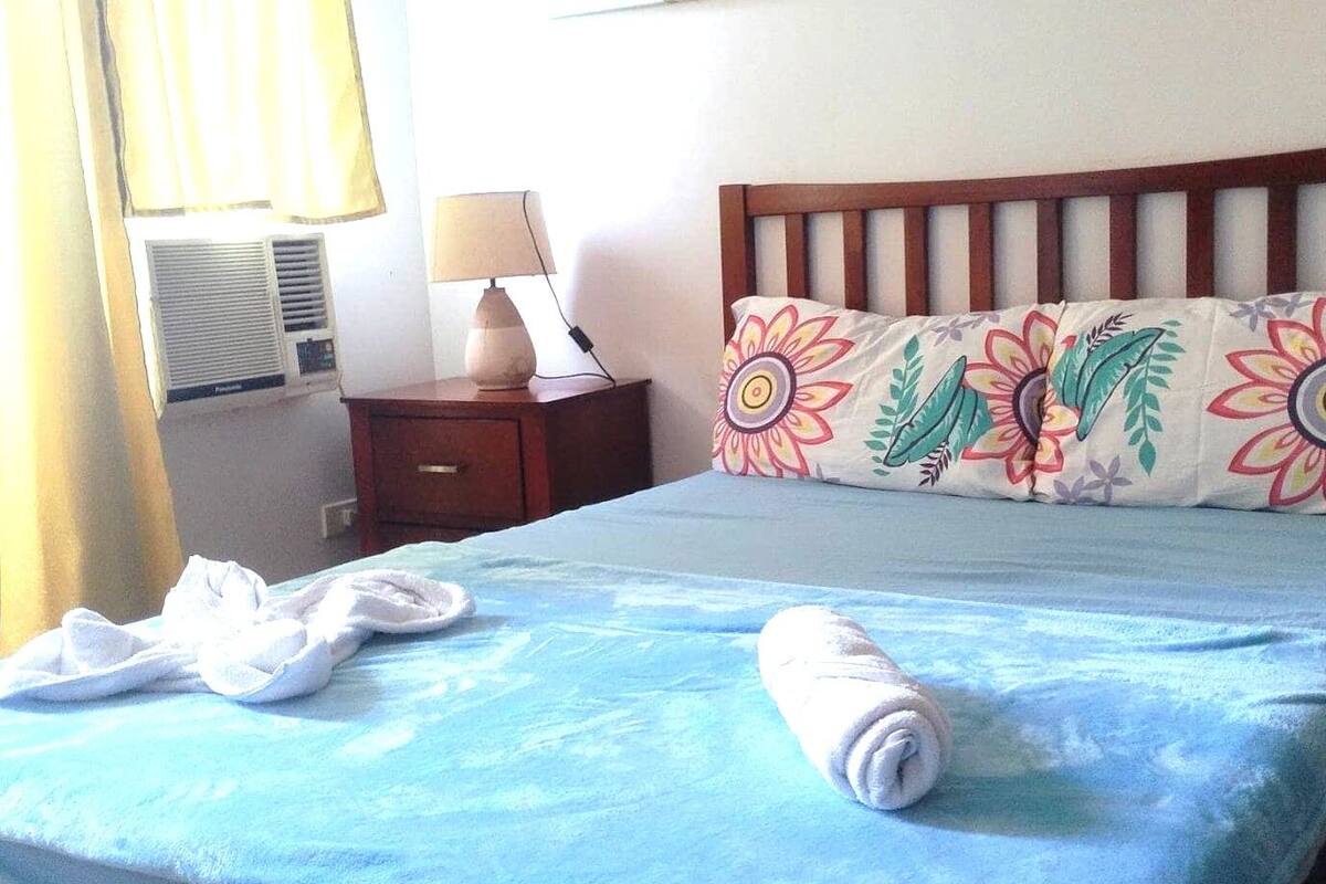 A hotel room with a bed, a nightstand, a lamp, a painting of a beach scene, and a window with curtains.