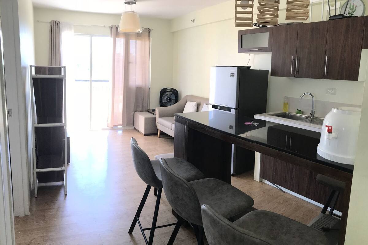 A modern kitchen with a refrigerator, sink, and bar stools. There is a dining area with a sofa and a window with curtains.