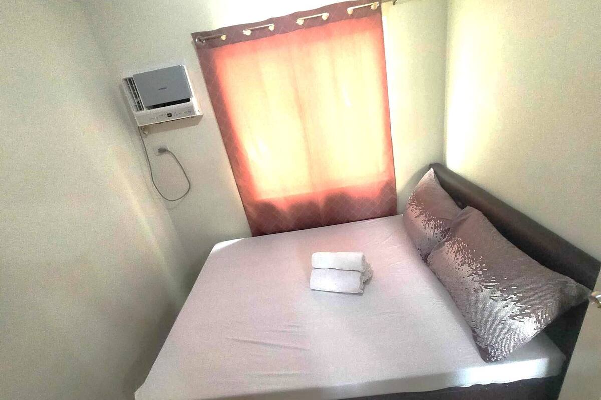 A single bed with a white mattress and pillows, a small air conditioning unit on the wall, and a window with a curtain.