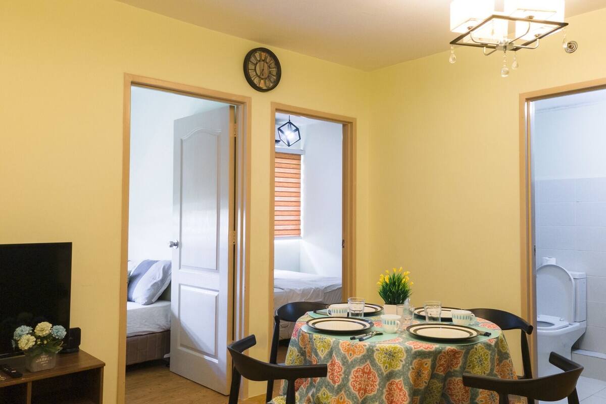 A compact room with a dining table set for two, a television on a stand, and a clock on the wall.