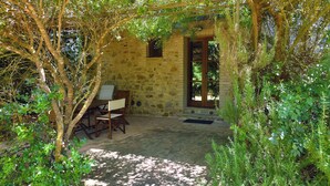 Edificio de piedra con puerta de madera y una pequeña ventana, rodeado de follaje y una mesa con sillas.