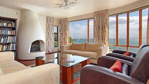 A spacious living room with a large window offering a view of the sea, a fireplace, and a bookshelf. - Private villa in Gordons Bay with ocean views and pool (Cape Town)