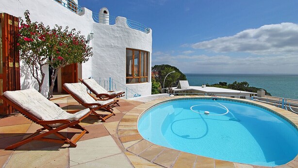 A pool area with lounge chairs, a clear blue pool, and a view of the sea. - Private villa in Gordons Bay with ocean views and pool (Cape Town)