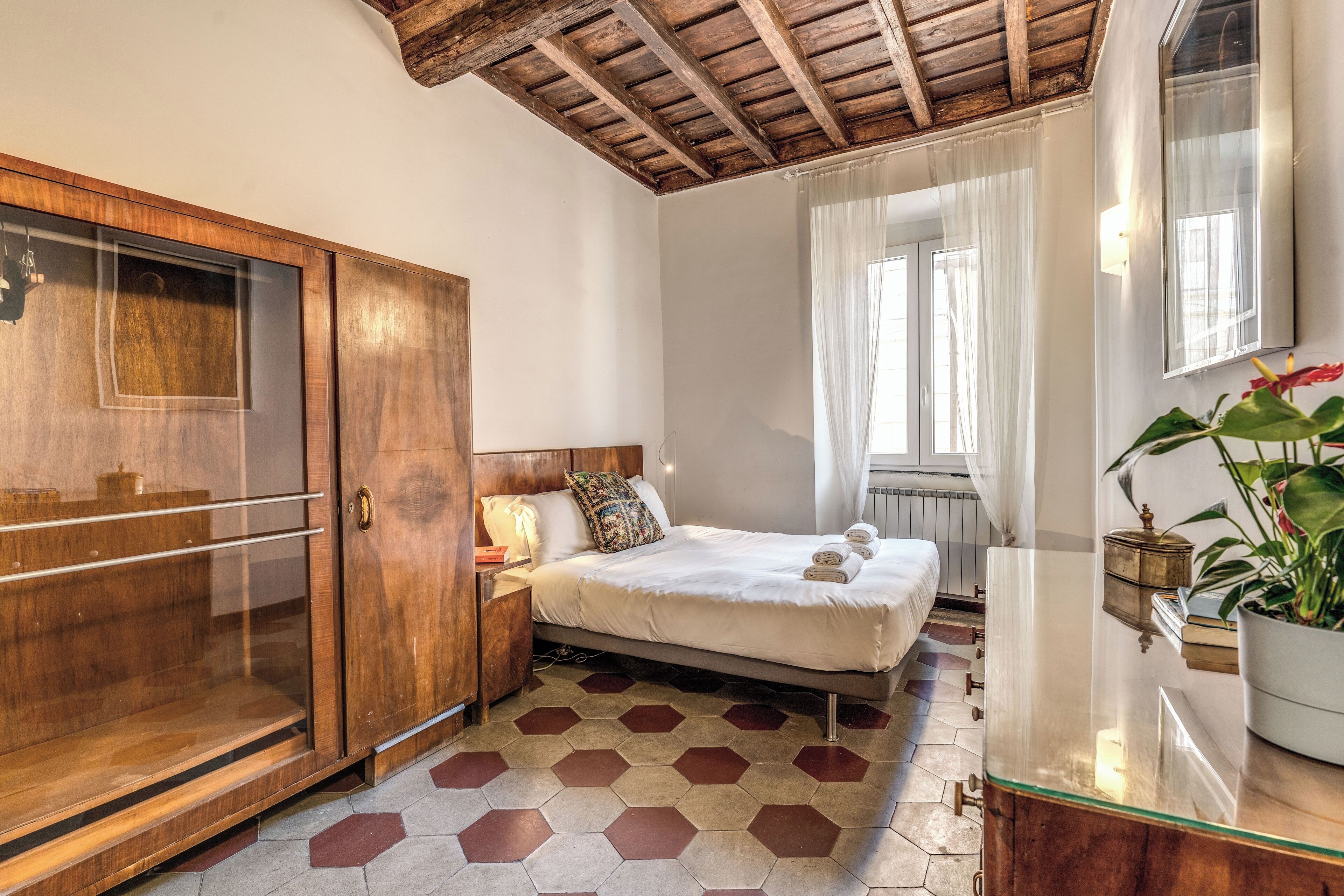 A bedroom with a wooden wardrobe, a bed with a white mattress, a bedside table with a lamp, and a window with curtains.