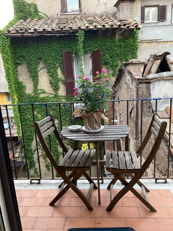 Un balcon avec une table ronde en bois et deux chaises en bois, un pot de fleurs, et une vue sur un immeuble dont les murs sont recouverts de lierre et qui possède des volets bruns.