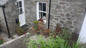 A stone house with a white door and windows, a small garden with flowers and a bench, and a sign reading "TLEOGGAN".