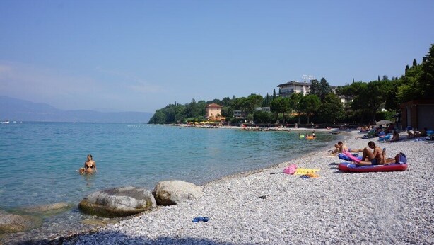 A pebbled beach with people sunbathing and swimming, a clear blue sky, and buildings in the background. - Lovely apartment on Lake Garda in residence with pool (San Felice del Benaco)