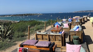 Een zitgedeelte aan het strand met houten tafels en banken, met uitzicht op de kust, boten en een heldere lucht.