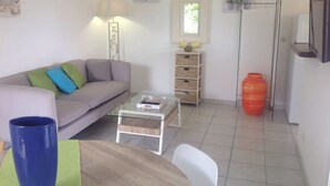 Una sala de estar con un sofá, una mesa de centro de vidrio, una silla blanca, una lámpara, un ventanal y un televisor.