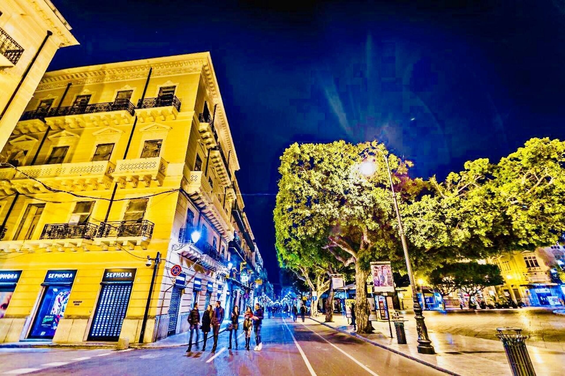 A nighttime street scene with a yellow building featuring balconies and storefronts, including a Sephora store.