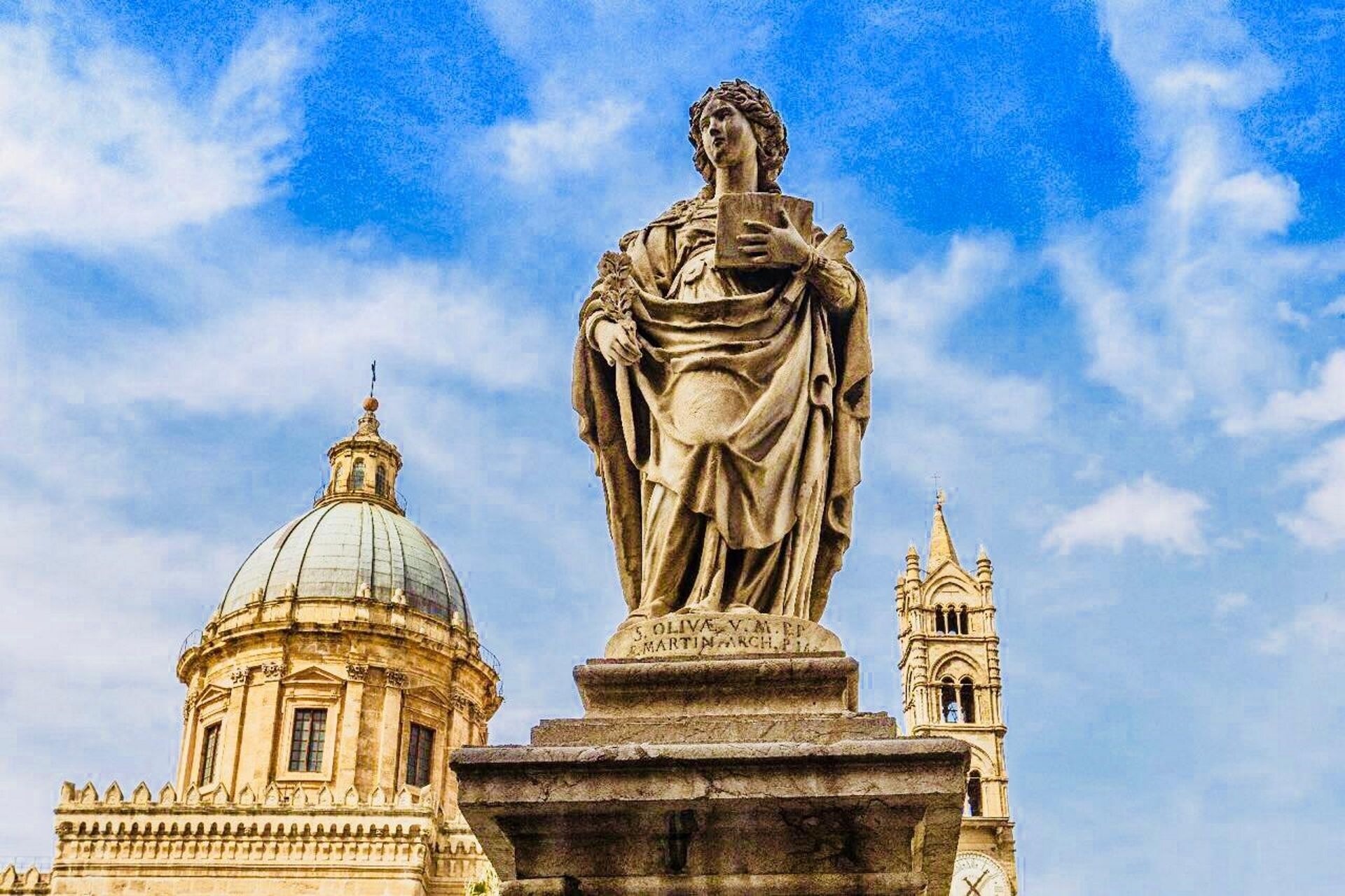 Una estatua de una mujer sosteniendo un libro, con una cúpula y un campanario al fondo.