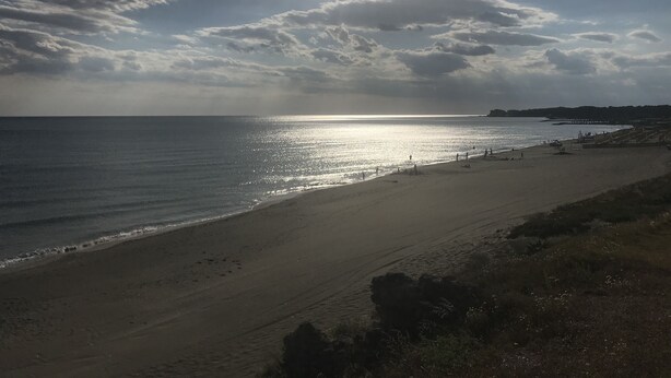 Una spiaggia con il sole che tramonta all'orizzonte, cielo sereno e una costa lontana.