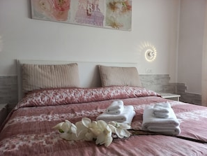 Un lit bien fait, recouvert d’une couvre-lit à motifs roses et blancs, avec deux oreillers beige et un luminaire fixé au mur.