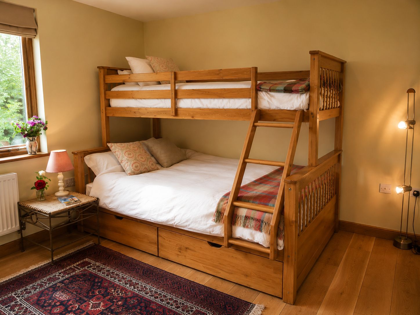 Un letto a castello con struttura in legno, un comodino con una lampada, un tappeto e una finestra che si affaccia sul verde.
