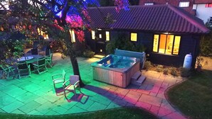 A backyard with a hot tub, patio furniture, and a house with illuminated windows.