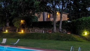 Piscina com espreguiçadeiras, um edifício de pedra ao fundo e vegetação ao redor.