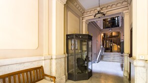 Ein mit Marmor ausgelegter Flur mit einer hölzernen Bank, einem gläsernen Aufzug und einer Treppe, die in eine obere Ebene führt.