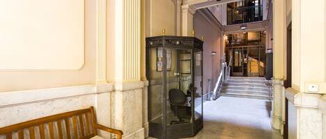 Un pasillo con suelo de mármol, un banco de madera, un ascensor de vidrio y una escalera que sube a un nivel superior.