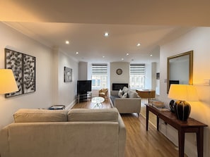 A modern living room with a sofa, TV, and a coffee table. There are windows with blinds, a clock on the wall, and a decorative wall piece.
