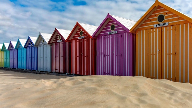 A row of colorful beach huts with names on signs, set against a sandy beach and a partly cloudy sky.