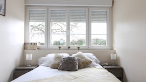 A neatly made bed with a plaid blanket, two bedside lamps, and a window with blinds.