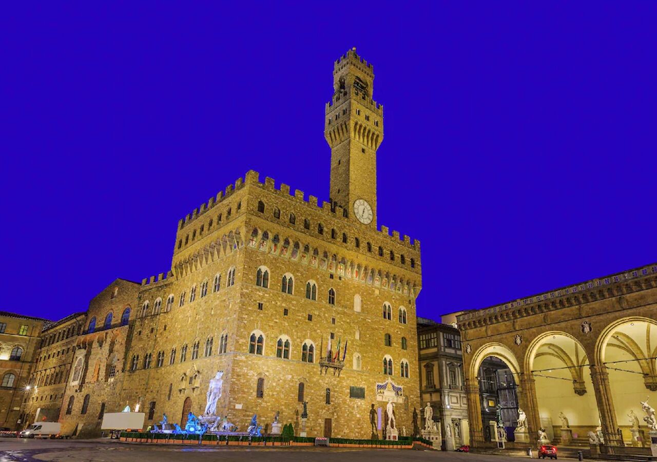 A historic building with a prominent tower and arched windows, featuring a clock and statues in the courtyard.