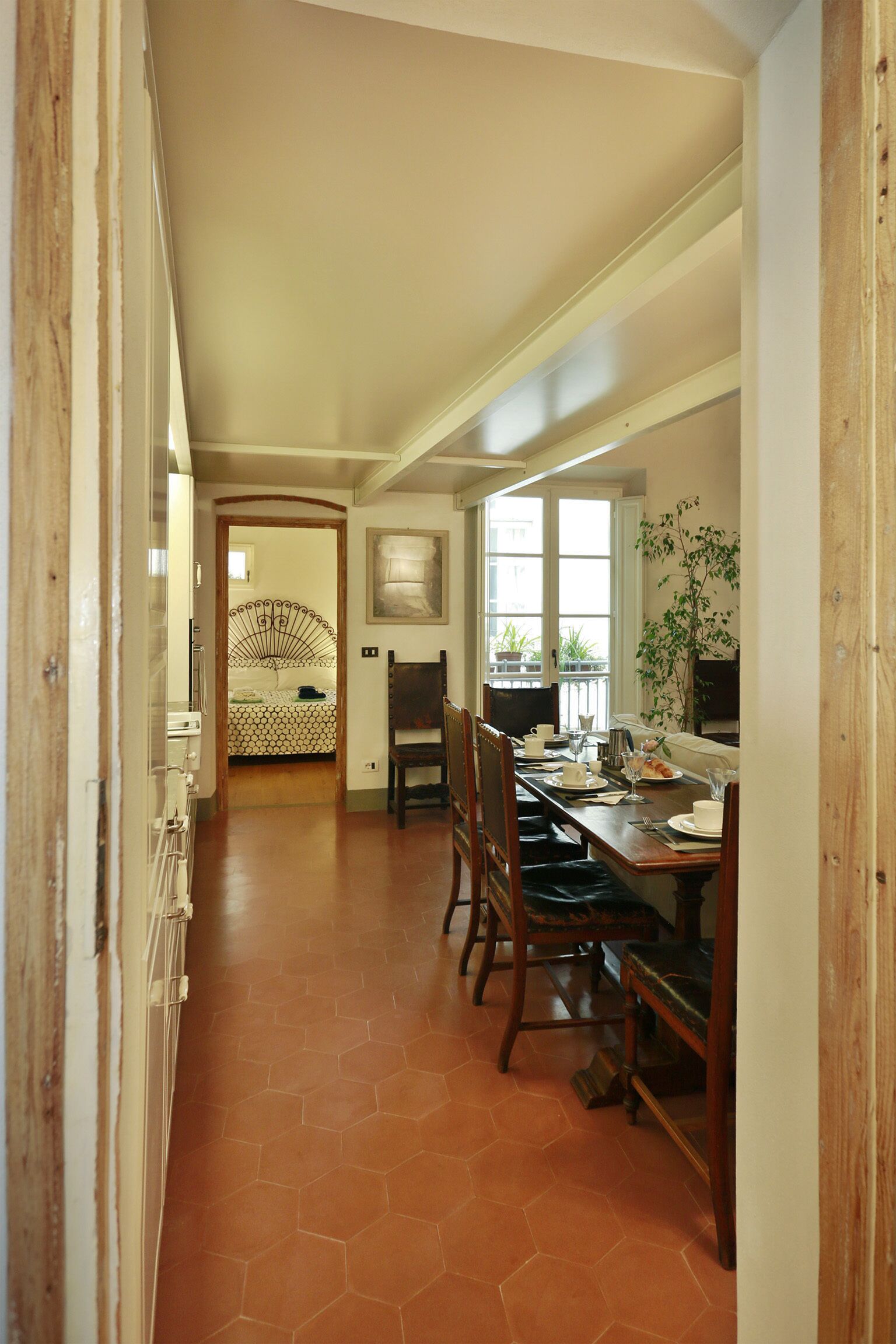 Un comedor con una mesa larga, sillas y vista a un dormitorio con cama y ventana.